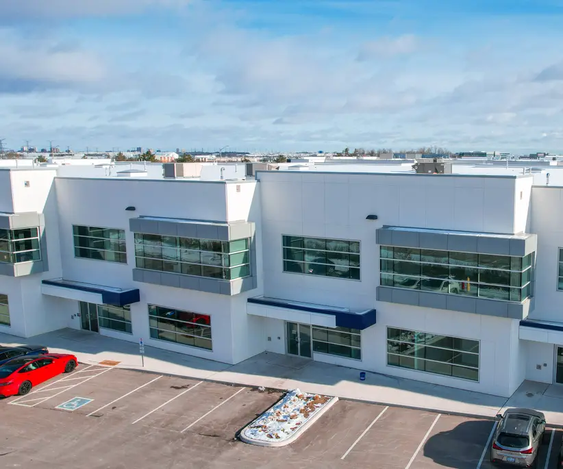Modern white commercial building with large windows, a red and a gray car in the parking lot, and a clear blue sky in the background.